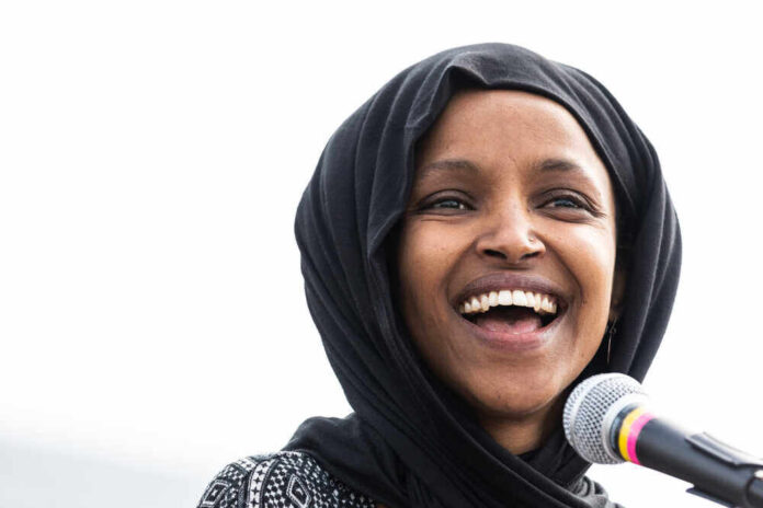 shutterstock_1369296272.jpg featured image Ilhan Omar with a black headscarf smiles near a microphone.