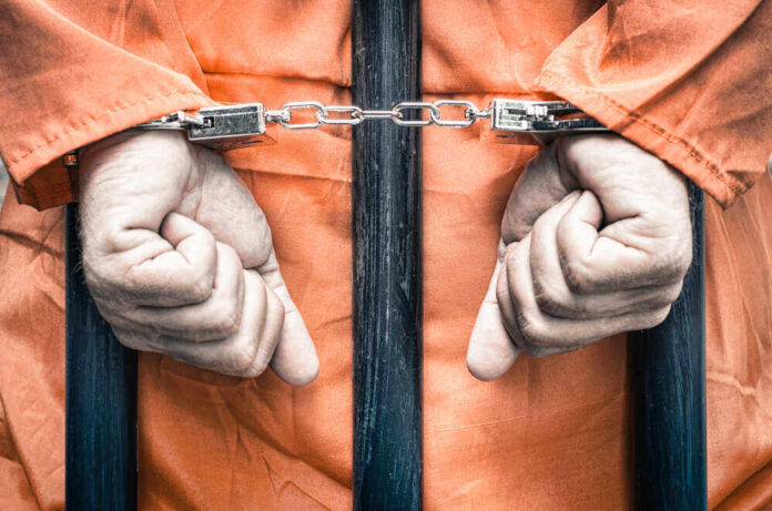 shutterstock_217094626.jpg featured image Handcuffed person in orange jumpsuit behind prison bars.