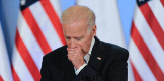 Joe Biden coughing in a suit standing in front of flags