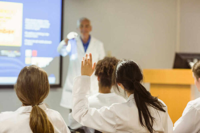 Students in lab coats raising hands in lecture.