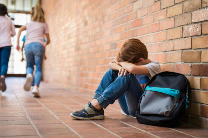 Sad child sitting against brick wall, kids running away