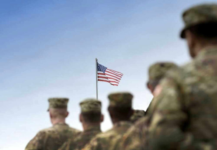 Soldiers in uniform facing American flag outdoors.