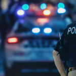 Full-Auto Ambush: Two Cops Barely Escape Execution Police officer stands near patrol car with lights flashing.