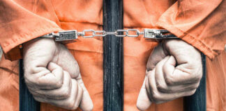 Handcuffed person in orange jumpsuit behind prison bars.