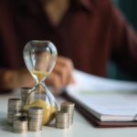 An hourglass next to stacks of coins on a desk with a person writing in the background