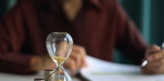 An hourglass next to stacks of coins on a desk with a person writing in the background