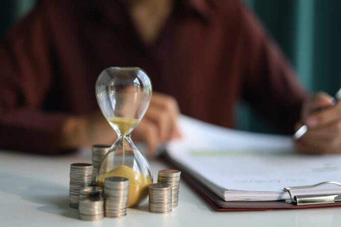 An hourglass next to stacks of coins on a desk with a person writing in the background