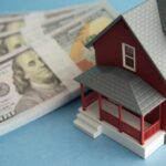 A model red house next to stacks of hundred-dollar bills