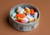 A bowl filled with various colorful pills and capsules next to a measuring tape