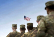 Soldiers in uniform facing American flag outdoors.