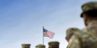 Soldiers in uniform facing American flag outdoors.