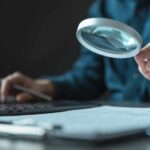Person using a magnifying glass while working on a laptop