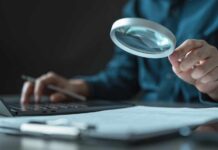 Banks Get Green Light From Trump To Check THIS Person using a magnifying glass while working on a laptop