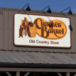 Cracker Barrel Axes THESE Restaurants — Why Now? Sign for Cracker Barrel Old Country Store featuring a man sitting on a barrel
