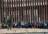 ICE Jail Emergency Ends In A Body Bag Border patrol officer watches group sitting by a fence.