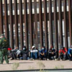 ICE Jail Emergency Ends In A Body Bag Border patrol officer watches group sitting by a fence.