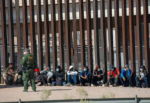 ICE Jail Emergency Ends In A Body Bag Border patrol officer watches group sitting by a fence.