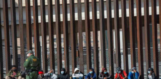 Border patrol officer watches group sitting by a fence.
