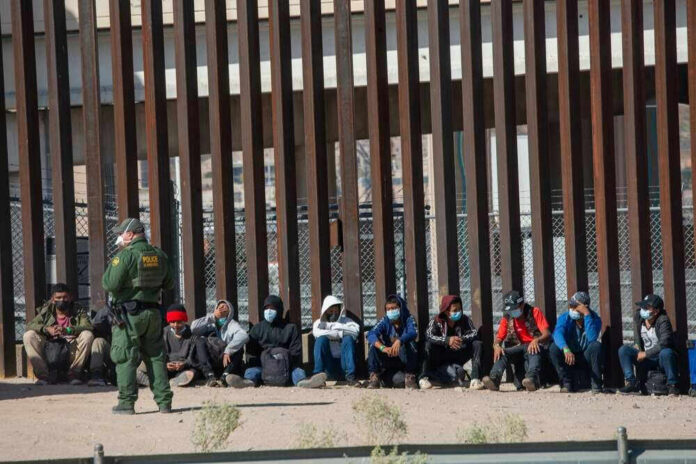 Border patrol officer watches group sitting by a fence.