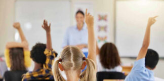 Children raising hands in classroom, teacher in background.