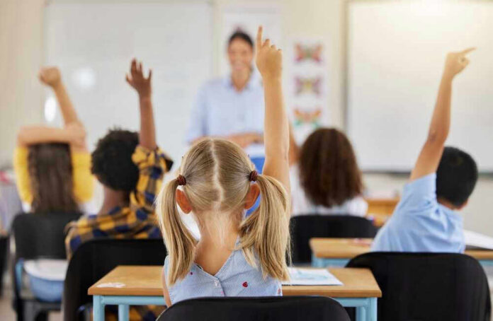 shutterstock_2282044199 Children raising hands in classroom, teacher in background.
