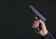 Armed Scout Nabbed At Trump Golf Hand holding a black semi-automatic pistol against a dark background