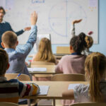 Students raising hands in a classroom with a teacher.