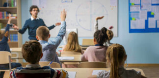 Students raising hands in a classroom with a teacher.