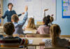 Students raising hands in a classroom with a teacher.