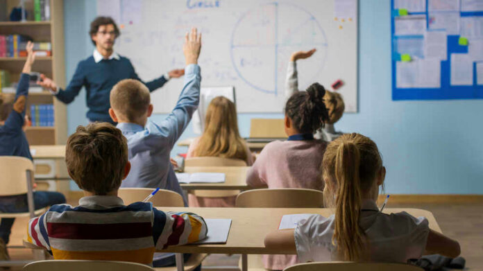 shutterstock_1767334112 Students raising hands in a classroom with a teacher.