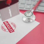 Deadly Measles Hits Blue State Again A stamp marking 'MEASLES ALERT' on a piece of paper next to a stethoscope and keyboard
