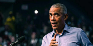Barack Obama in blue shirt speaking at podium with microphone.