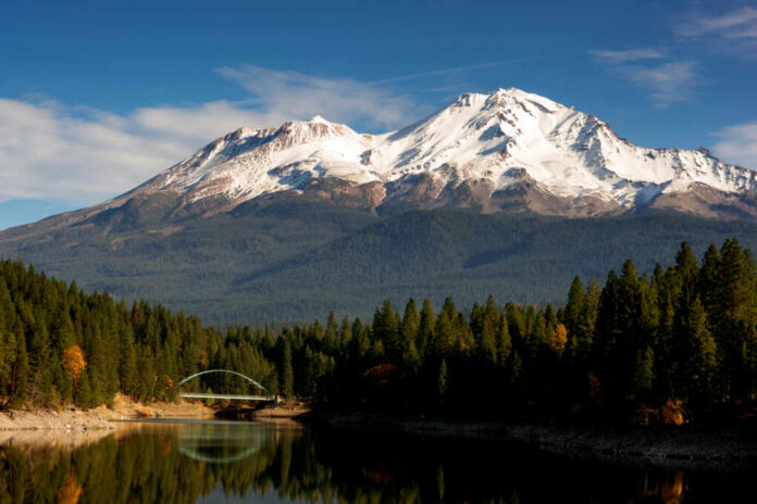 Mount,Shasta,Standing,Above,A,Lake,With,The,Name,Siskiyou Snow-capped mountain above a forested lake with a small arched bridge