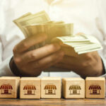 Bill Shock Spreads – Is Water Next? Person counting cash with wooden blocks depicting store icons