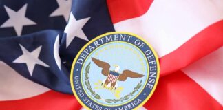 Soldiers in uniform facing American flag outdoors.